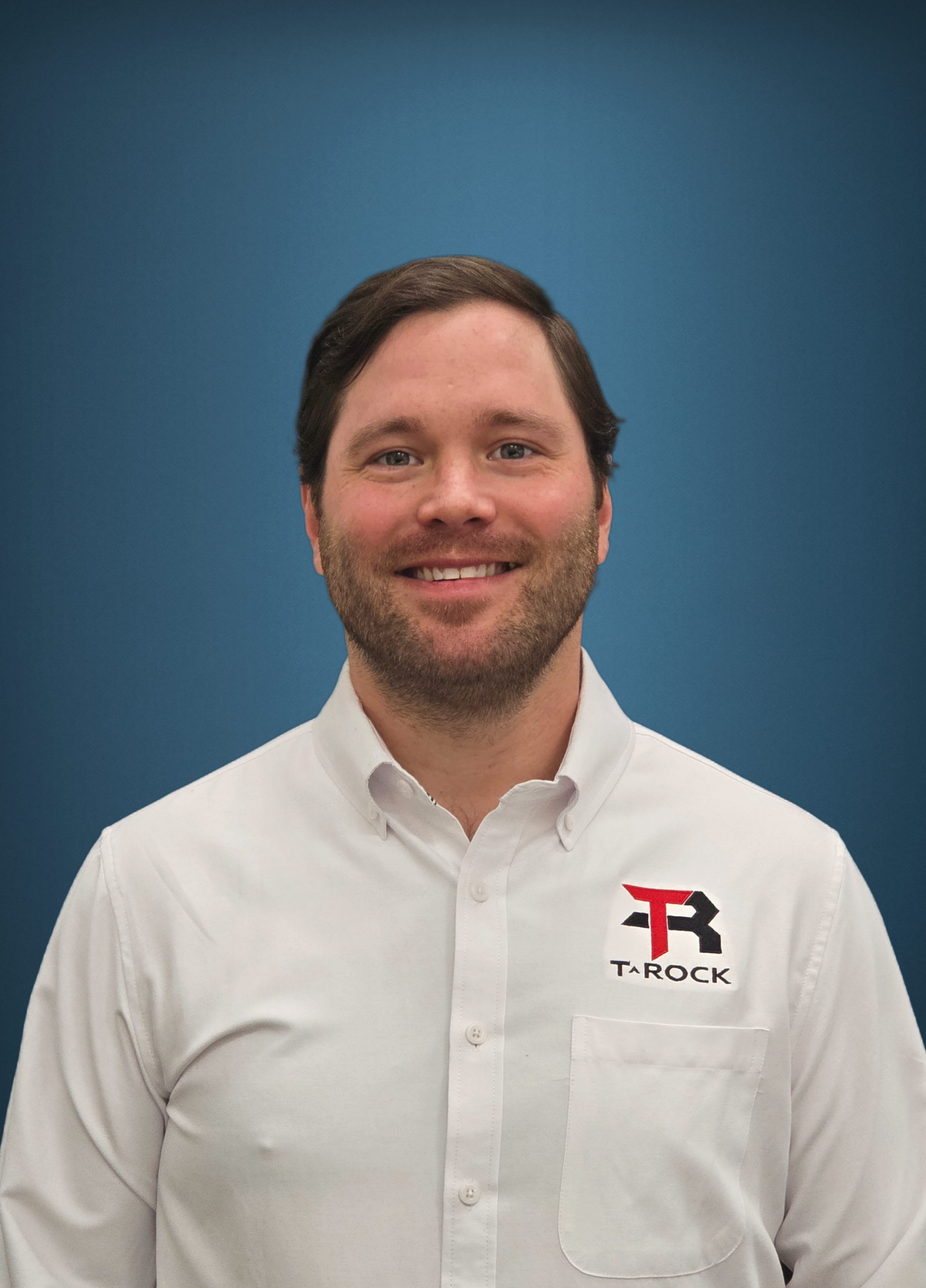 Portrait of a man with short brown hair and light stubble, smiling, wearing a white button-down shirt with a TAROCK logo on the left chest against a blue gradient background.