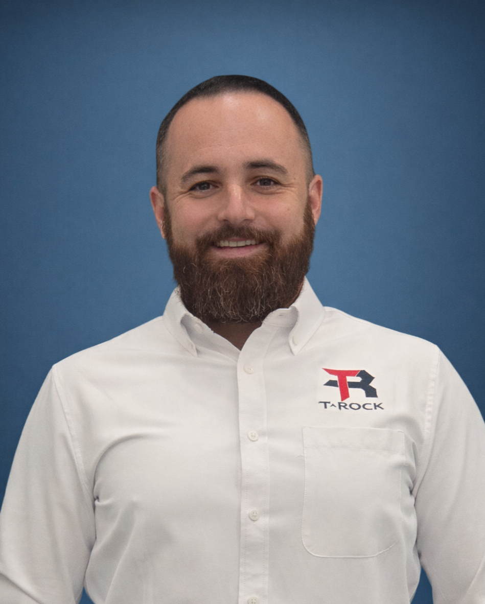 Smiling man with a beard in a white button-down shirt featuring a red and black T-Rock logo, standing against a blue backdrop.