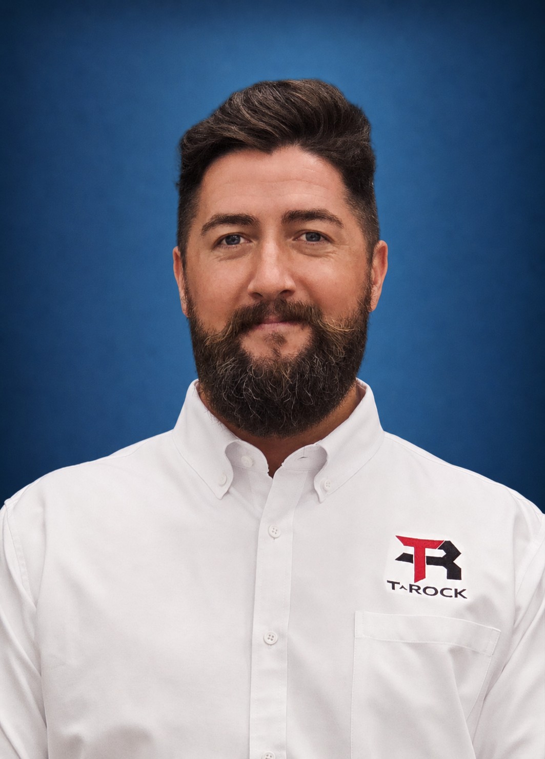 Portrait of a bearded man with dark hair in a white shirt featuring a Tarock logo on the chest against a blue background.
