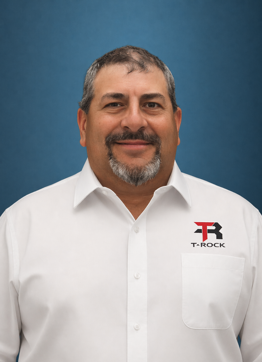 Portrait of a middle-aged man with short gray hair and a goatee, wearing a white shirt with a TAROCK logo on the chest against a blue background.