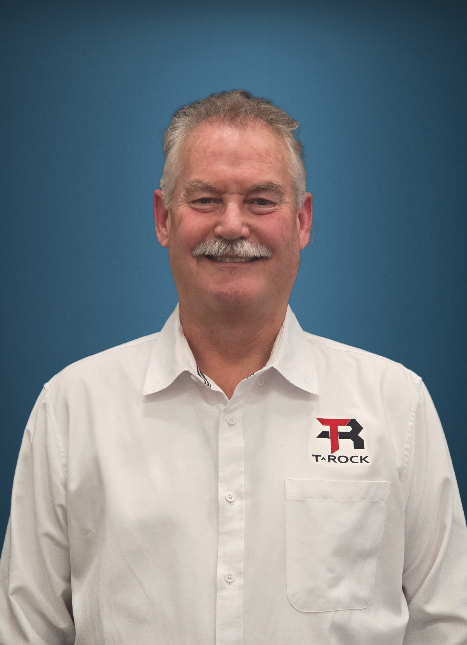 Smiling man in a white button-down shirt with a TAROCK logo on the left chest against a blue background.