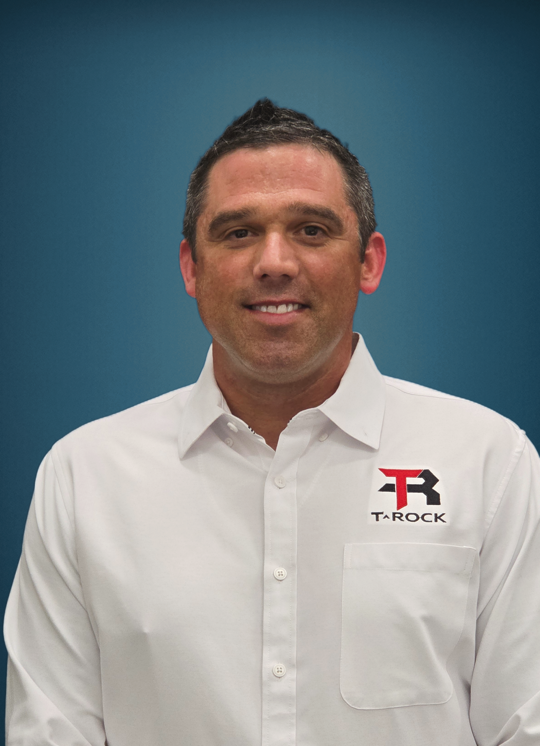 Portrait of a man in a white button-down shirt with a T-ROCK logo on the left chest against a blue background, smiling subtly.