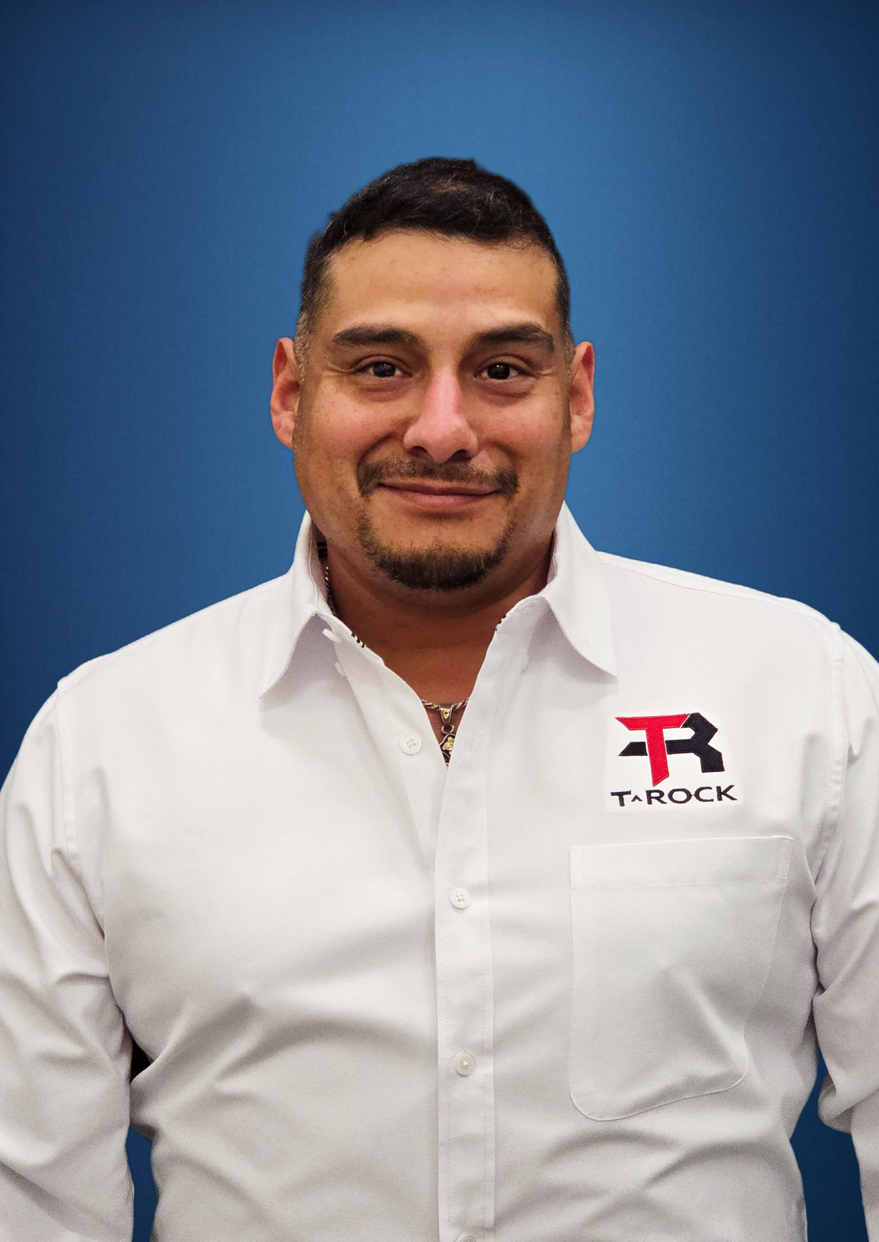 Man in a white shirt with a T-ROCK logo on the left chest, standing against a blue background.